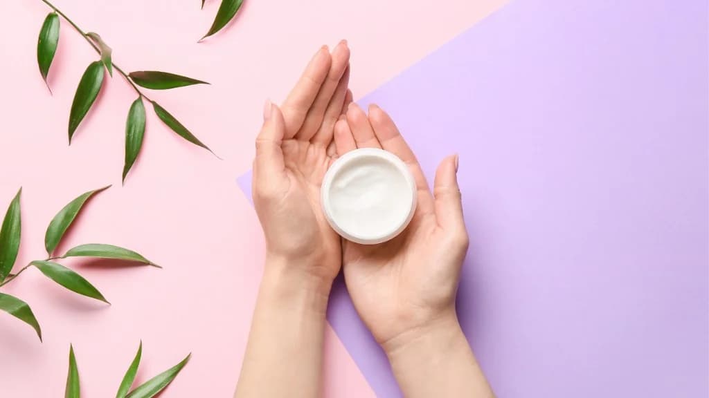 Hands holding a moisturizer jar over a pink and lavender background during a daily skincare ritual